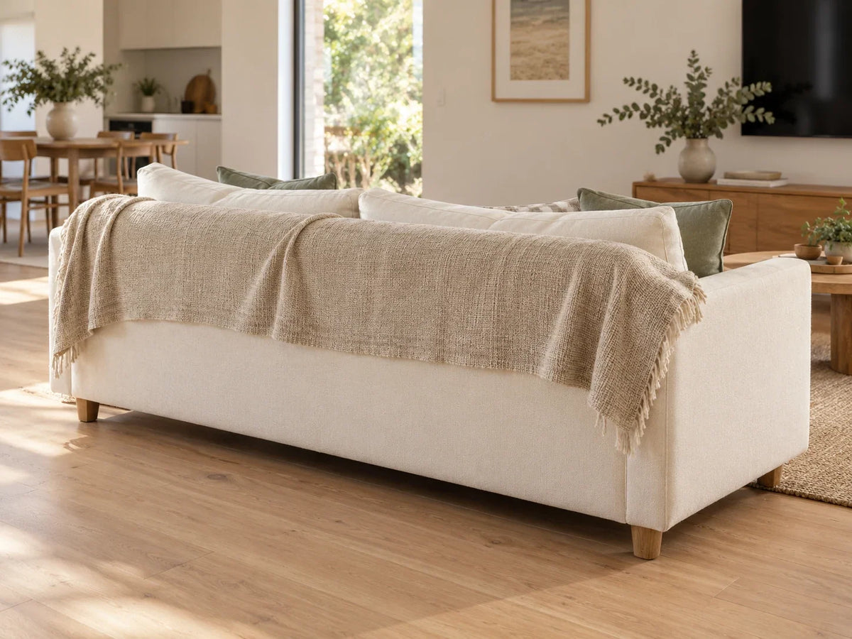 Cream sofa viewed from behind in a light open-plan room, with an oat textile draped over its upper back and lower upholstery visible.