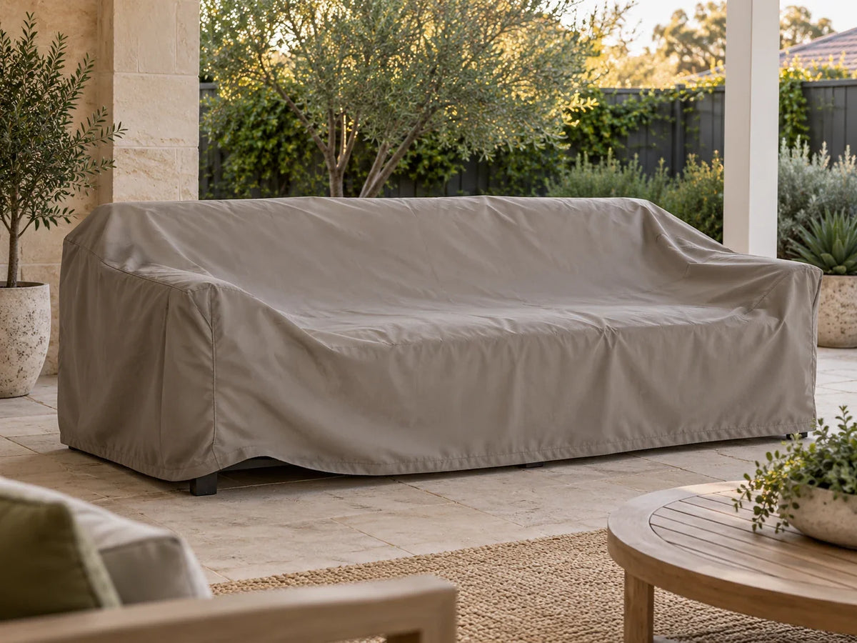 A plain taupe cover over an outdoor sofa on a calm patio, with part of the front hem slightly raised.