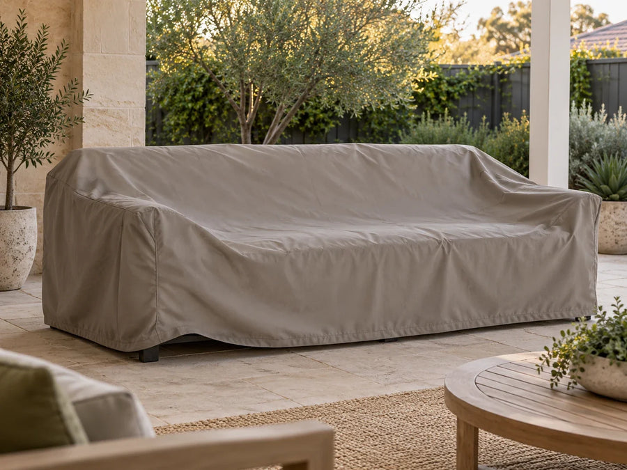 A plain taupe cover over an outdoor sofa on a calm patio, with part of the front hem slightly raised.