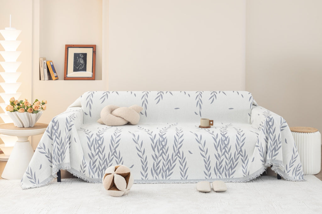Oliva throw with grey leafy branches draped over a sofa with styled cushions and fringed edges
